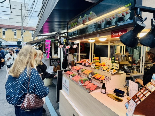築地場外市場商店街で肉の串焼きとハンバーガーを注文する外国人